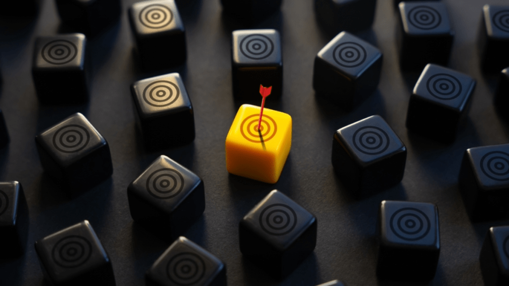 3D dice, all black, one yellow die in the center with a red arrow hitting a bullseye on the front face of the die
